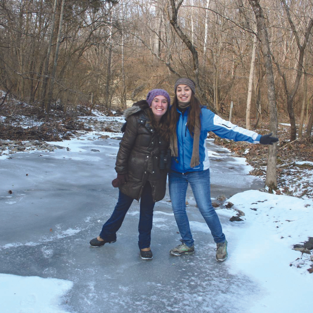 Two young women stand in winter gear on an icy stream with their arms around each other. 